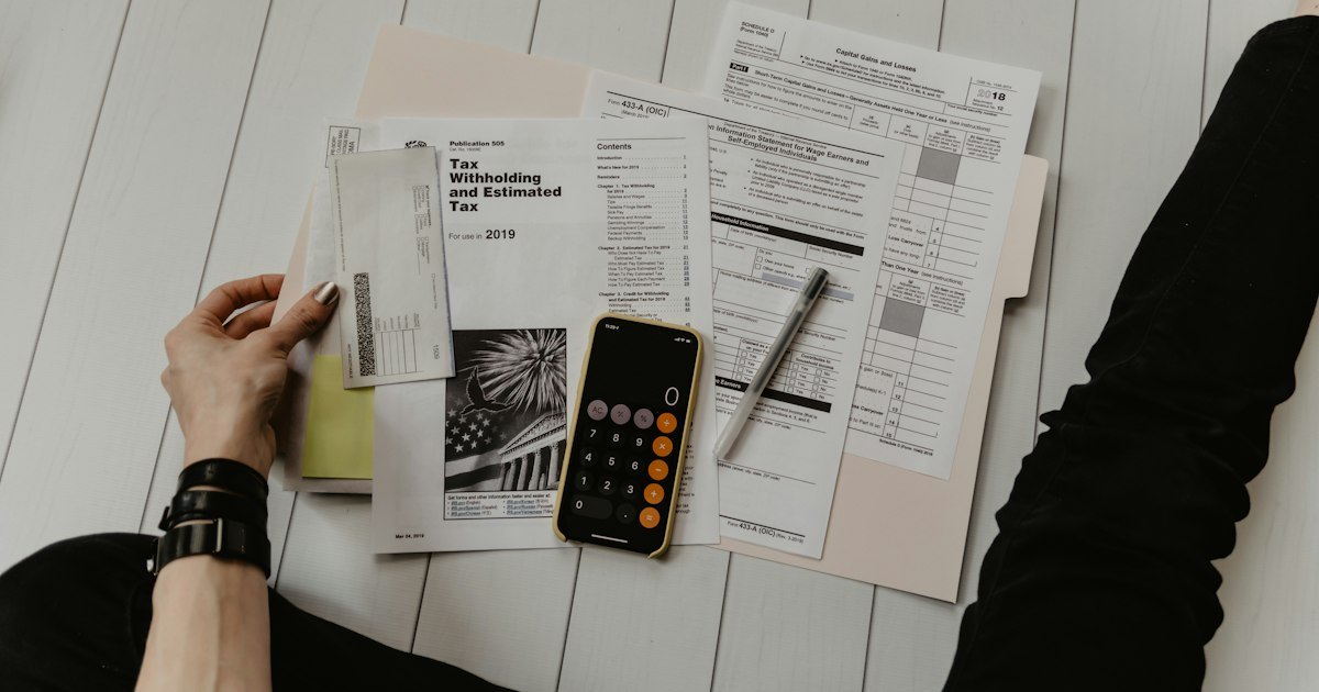 Hands filling in printed tax forms on a white surface with a calculator and paperwork