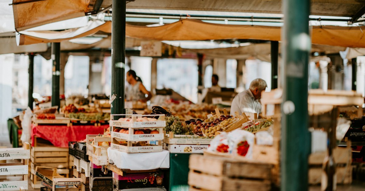 A covered market aisle with stalls stacked with crates of fruit, vegetables and packaged goods